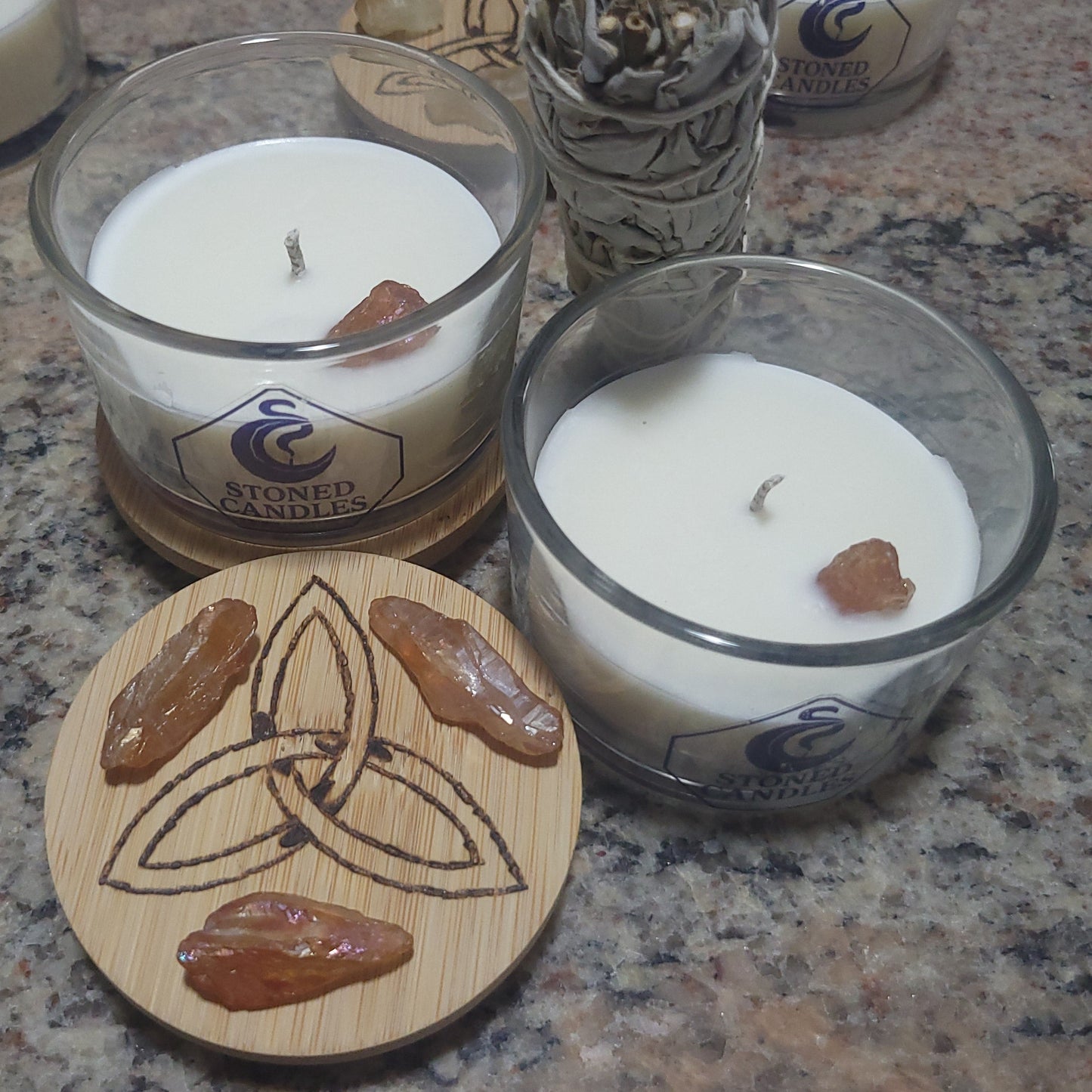 Glass candle containers holding crystal-embedded candles and wooden lids featuring Celtic knot designs on a granite surface.
